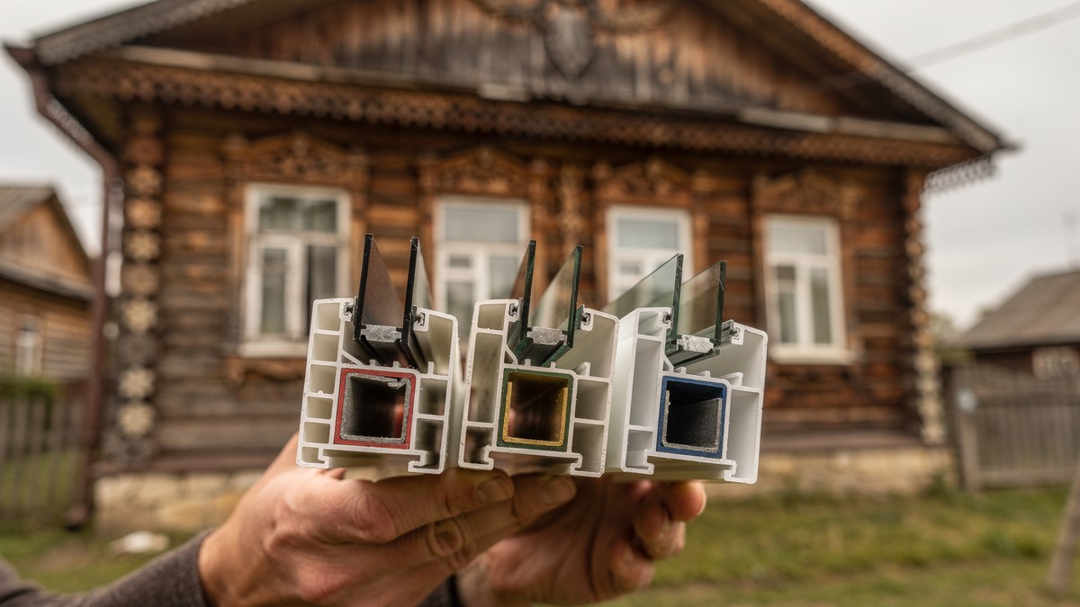 Монтажник держит образцы профилей Rehau, KBE и Veka на фоне деревянного дома в Тверицкой слободе
